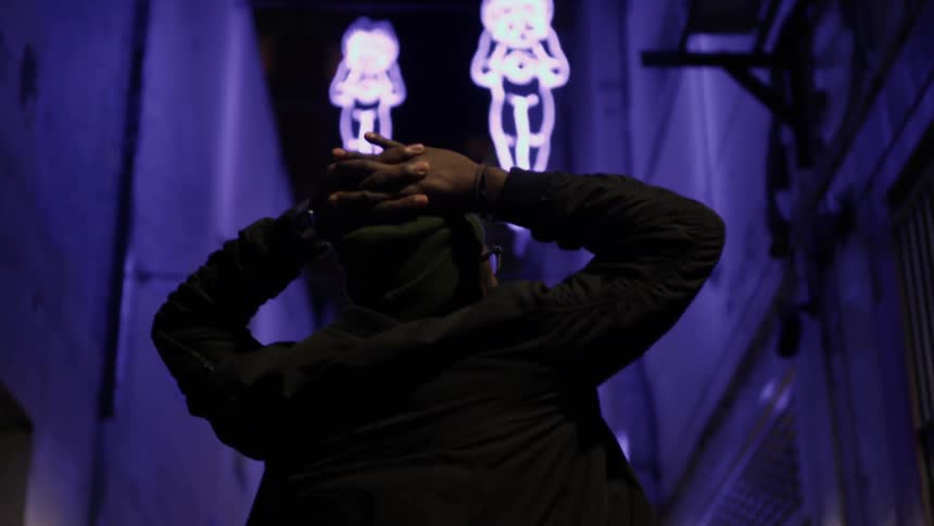 A man with his hands over his head looks up at neon lights