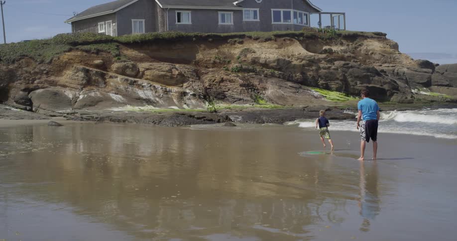 A man helps a boy ride a board across the wet beach