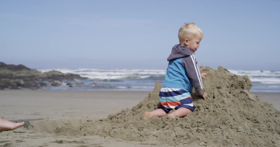 A man shoveling sand as a small boy leans against the sand pile and others are seen off in the distance walking along the beach