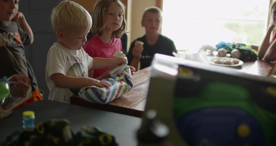 A woman and several children sitting at a table as a young boy opens a gift