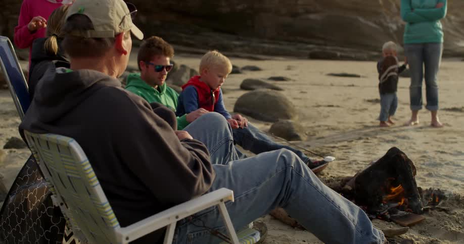 People are sitting on the beach roasting marshmallows on a campfire