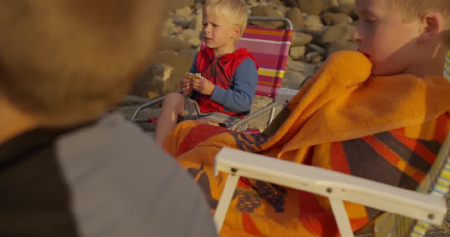 A couple of adults and some children sitting around a beach fire while one adult cooks a hot dog in the fire, one child eats and another sits with a towel wrapped tightly around him