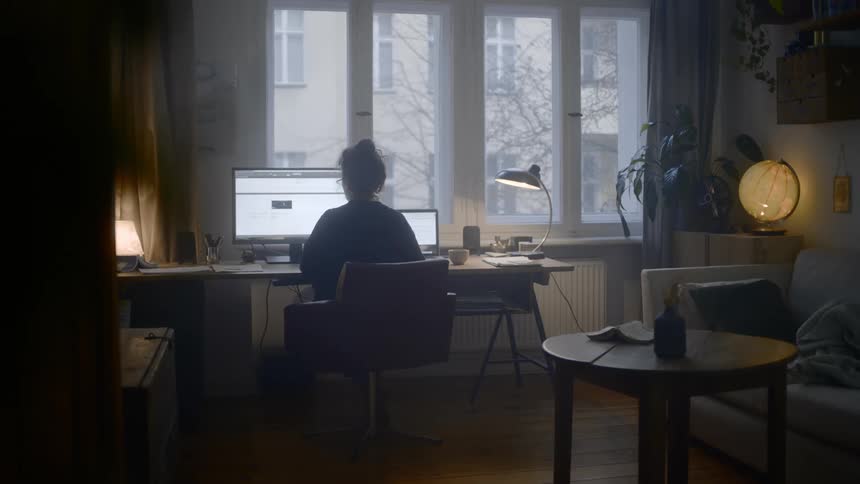 A woman sits at a desk with a large monitor and laptop and drinks from a small mug