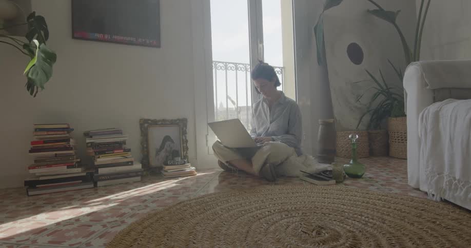 A woman sits on the floor of her living room and uses a laptop