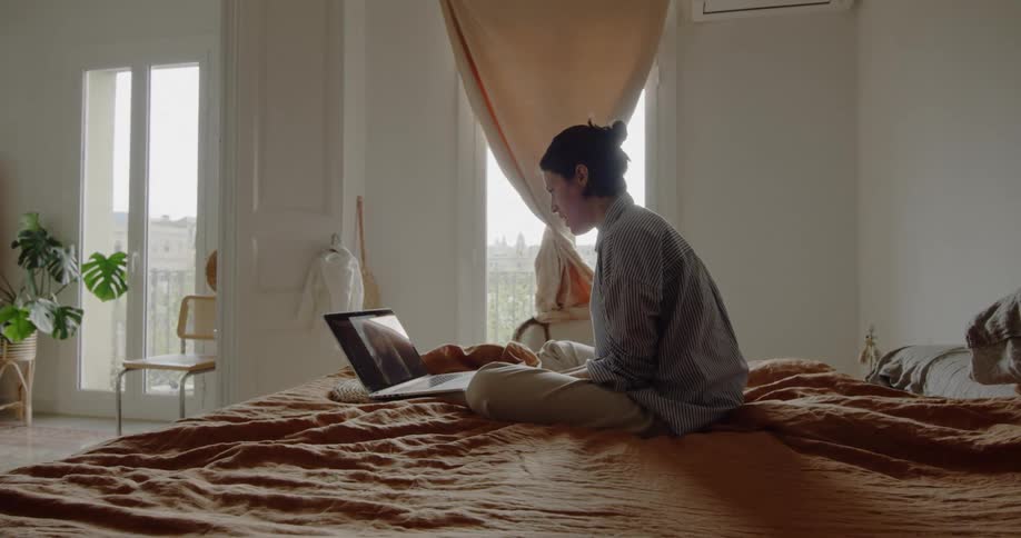 A white woman sits on a bed while talking to a person through a laptop
