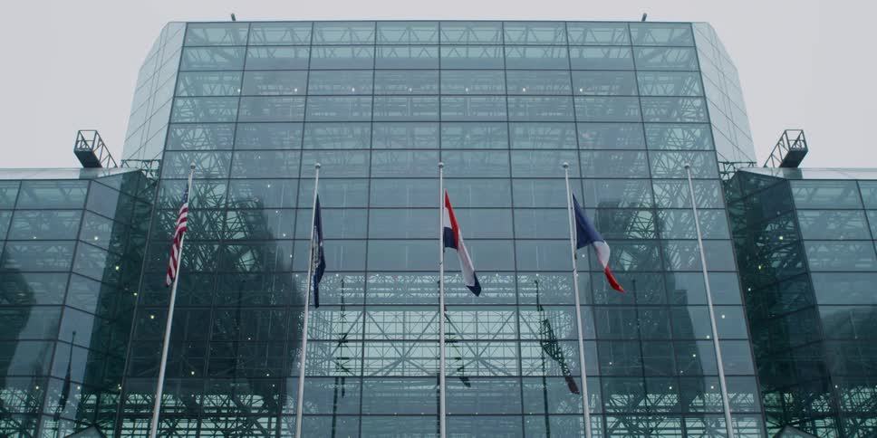 Five flagpoles stand in front of a glass building with metal framework visible