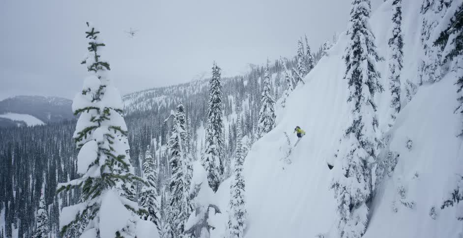 A skier jumps down a snowy cliff while a drone flies overhead