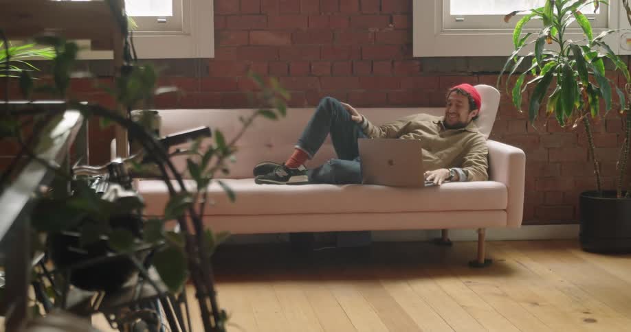 A man lies on his couch with his laptop open as he talks with someone on it