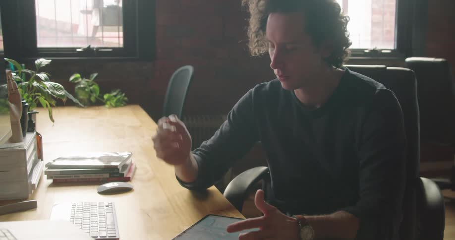 Man works on tablet while video conferencing at computer at desk in office