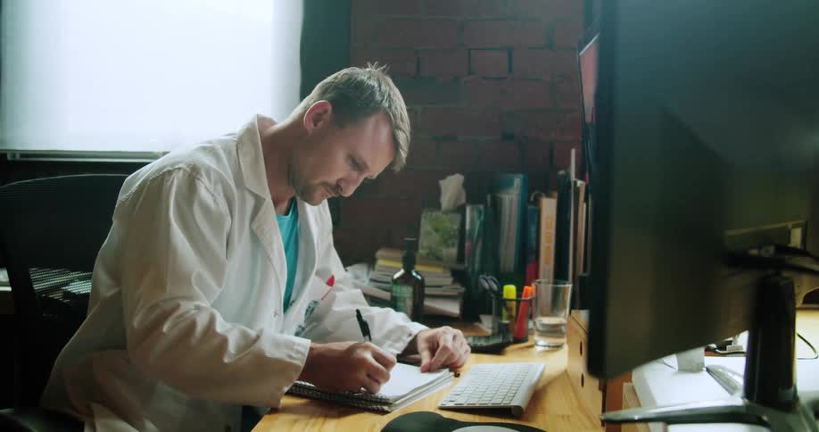A doctor writes in a notebook while looking at a computer screen