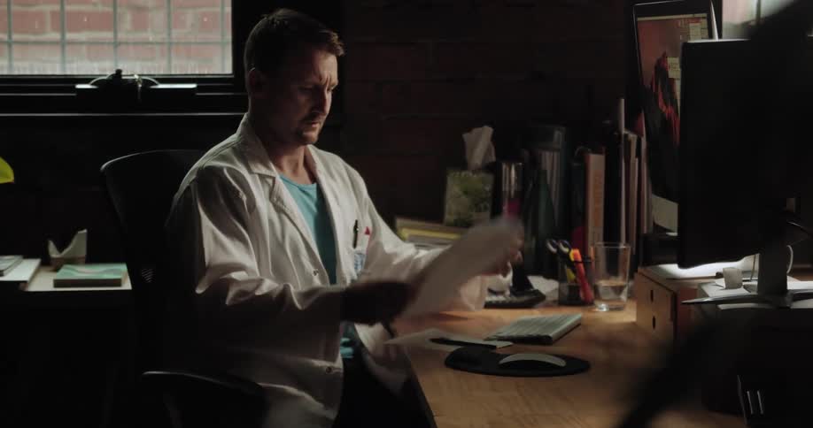 Doctor sits at desk reading document and typing on computer