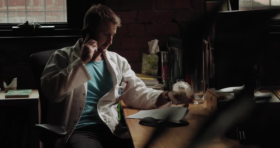 A man sits at his desk and looks at his computer and a piece of paper while talking on the phone