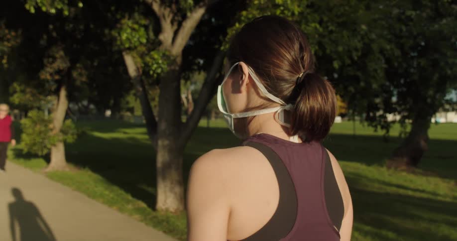 Female walks along the sidewalk next to the park filled with trees while wearing a face mask and approaching an older couple who is also walking