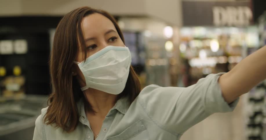 A woman in face mask and latex gloves talking tissue from a shelf and moving it to her shopping cart