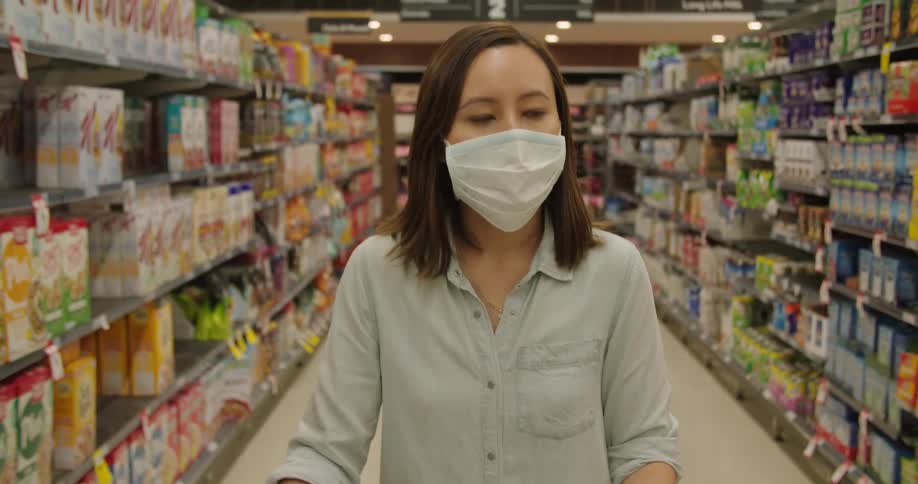 Asian woman wearing a face mask drives shopping trolley through food department in the supermarket