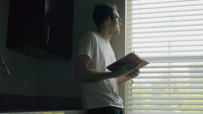 A man holding a book leans against the counter and looks out the window then goes back to reading