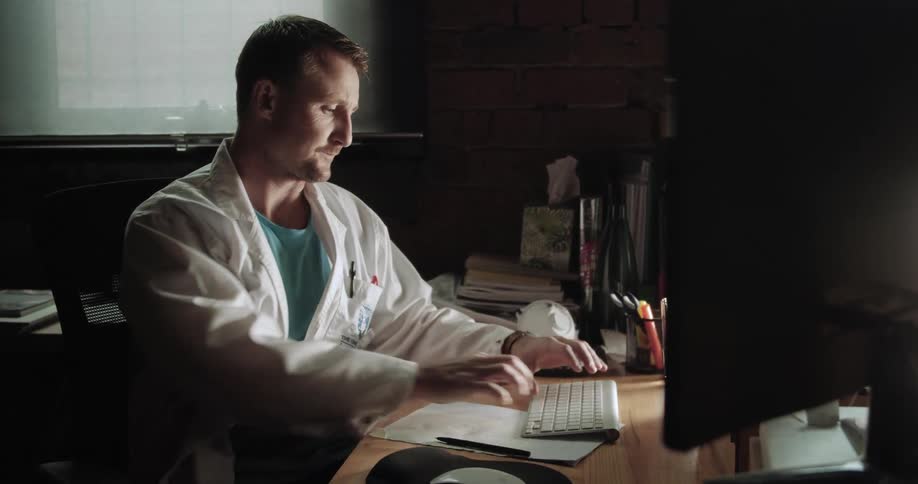 Doctor sitting at the table uses desktop and takes a note in the office
