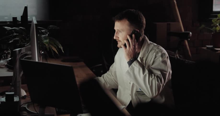 A man wearing a doctors coat talks on a cell phone while doing work on a computer
