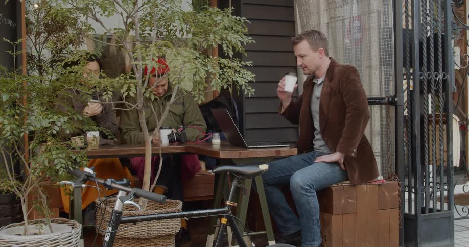 A man types on a laptop while sitting at a table drinking coffee with friends