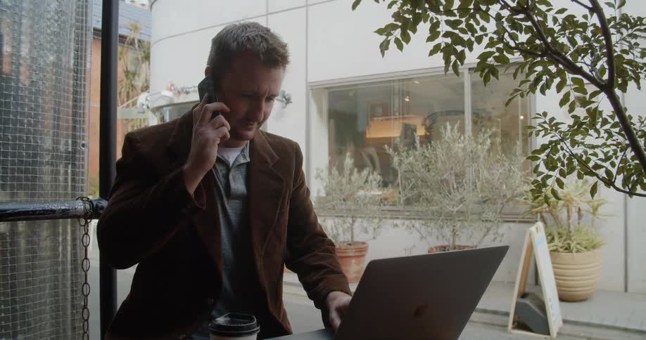 Man sitting at the table uses a laptop and talks to someone on the smartphone in the cafe