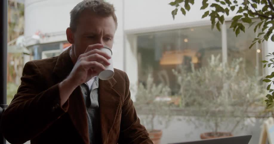 A man sitting at a table sipping on tea typing on his laptop