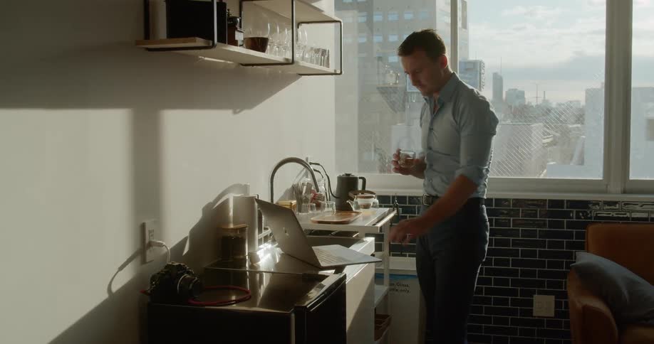 A man in a kitchen typing in a laptop and drinking a drink