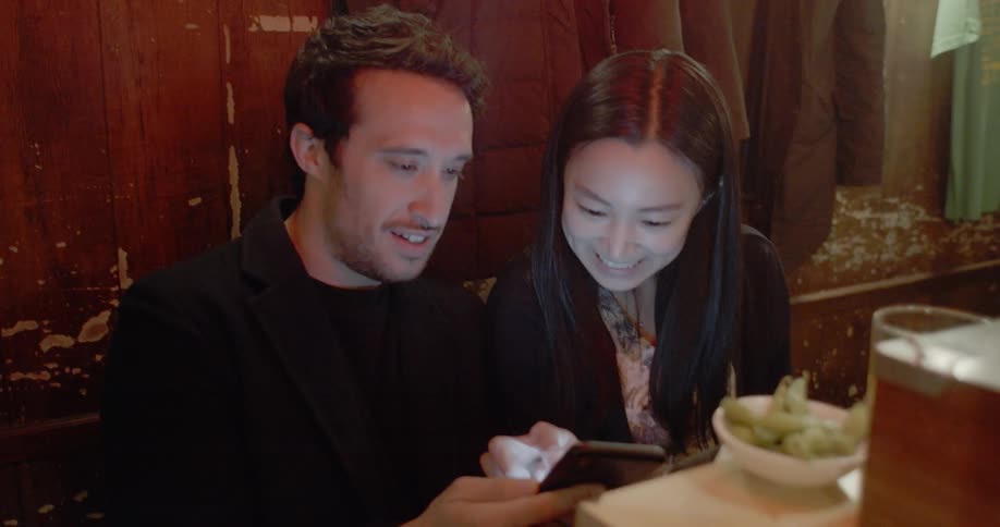 A man and woman sit at a restaurant and scroll through a cell phone and laugh