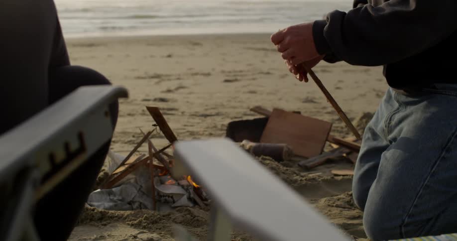 A man is kneeling down on the beach starting a fire