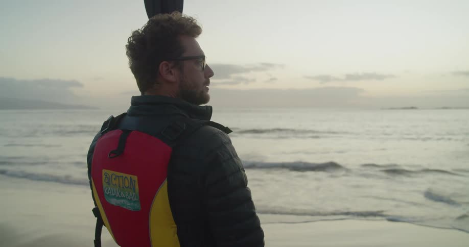 A man stands beside a kayak and looks out at the water