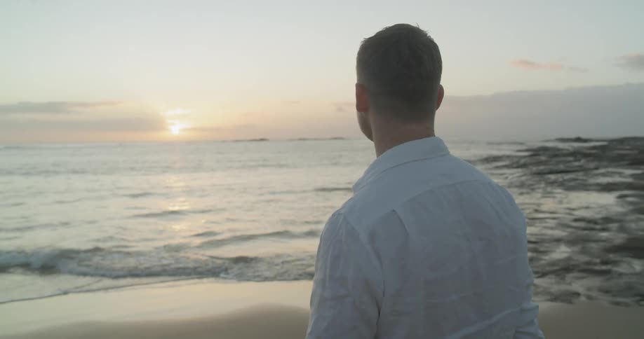 Man standing on the sand beach pets his dog at sunset next to the ocean