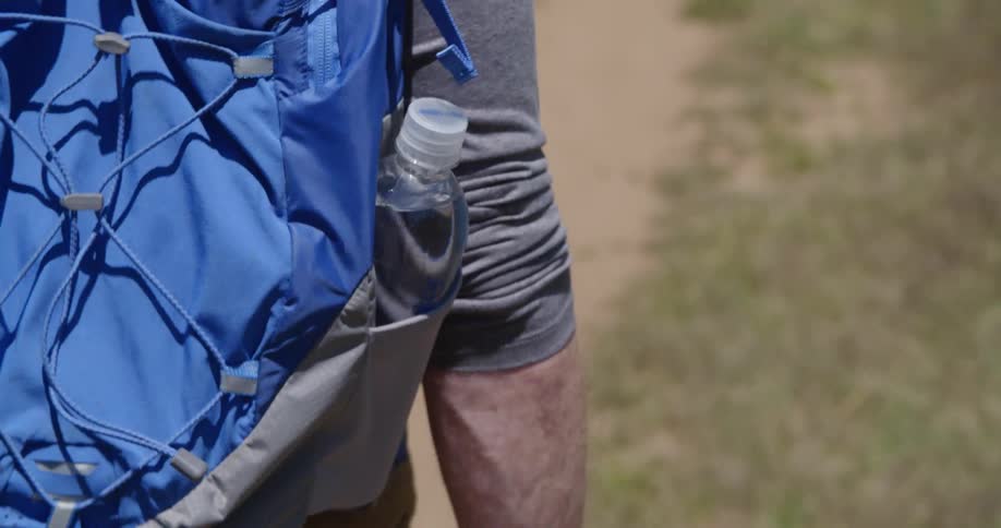A water bottle in the outer pocket of a backpack worn by a man walking on a trail on a sunny day
