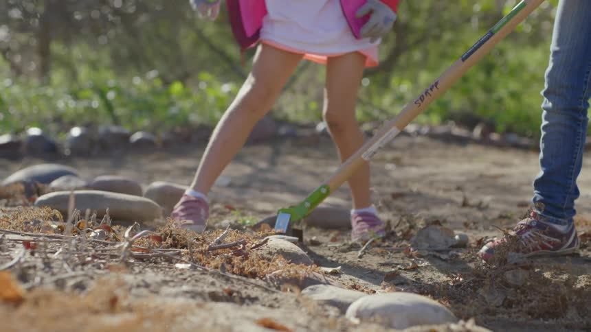 A little girl pats the ground as an adult rubs a tool on it