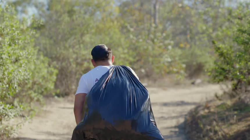 Man walks down dirt road carrying full plastic garbage bag over his shoulder