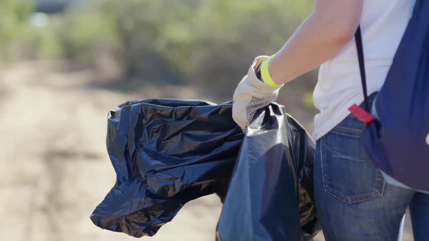 Person walks down dirt road carrying black plastic trash bag