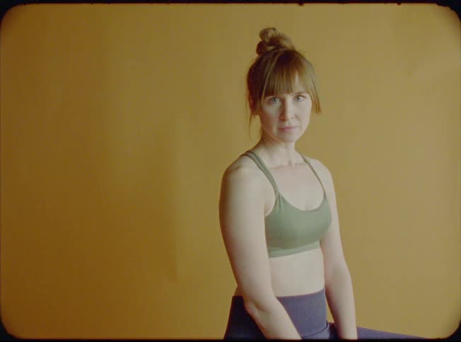 Blonde woman sitting on the stool looks at the camera with yellow wall in the background
