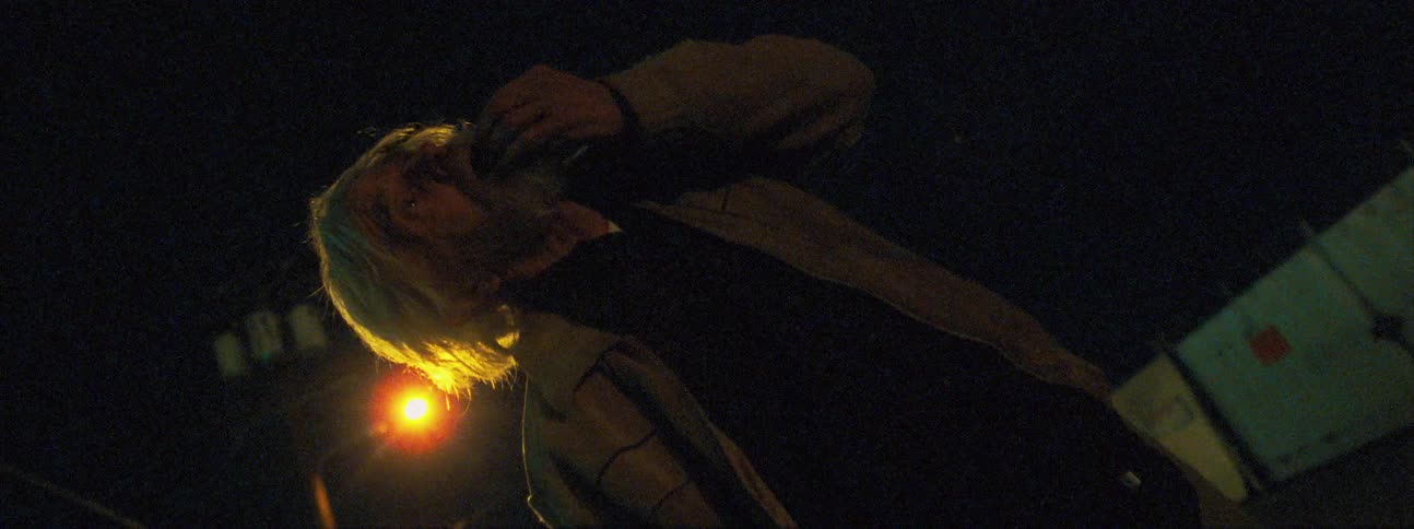 An elderly man shakes and touches his face while standing outside at night