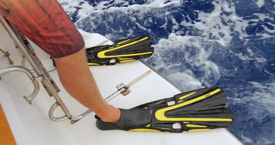 The bottom half of a man wearing scuba flippers and shorts are seen on the edge of a speeding boat