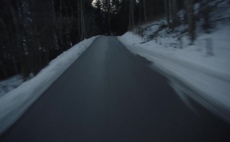 A narrow curving road through the trees with snow covered fields and slopes