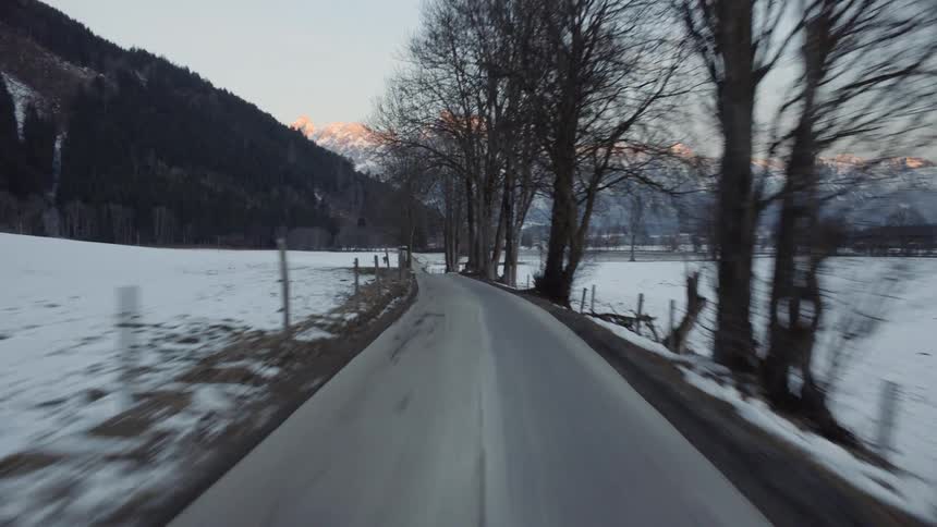 A narrow country road cuts through snow covered fields heading toward mountains