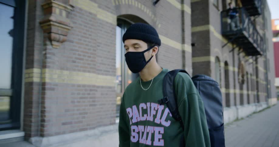 A college student wearing a mask and carrying a backpack walks past his empty school buildings