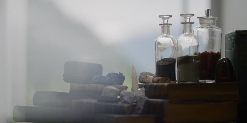 A collection of rocks and jars sit on antique books near a window with a hillside view