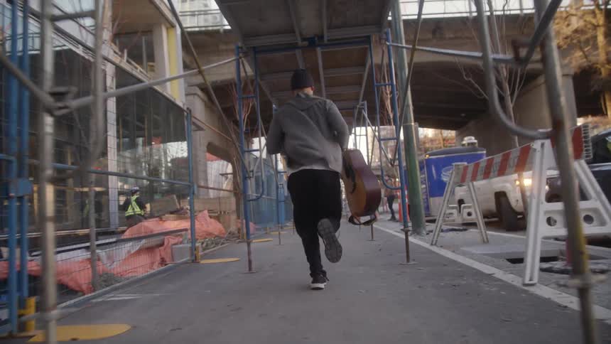 Man carrying a guitar runs along the pavement next to the road in the city
