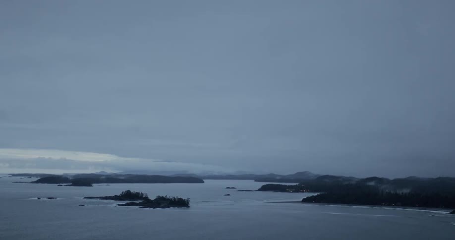 Lights glow onshore as ocean waves break against rocks and small islands on a gray cloudy day