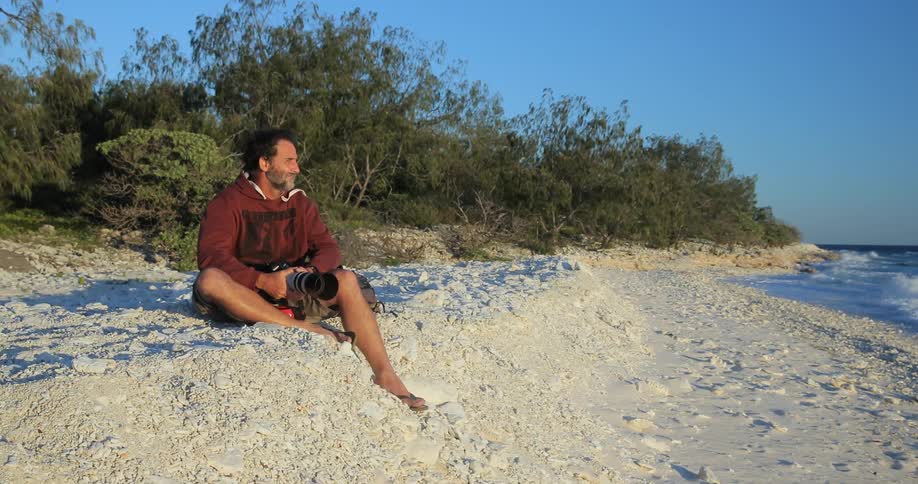 A man sits on a beach holding a large camera