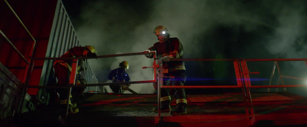 Firefighters standing on a metal structure emitting smoke as sirens flash