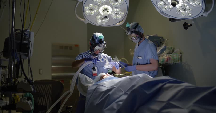 An anesthesiologist puts a mask on a patient while a surgeon prepares for surgery
