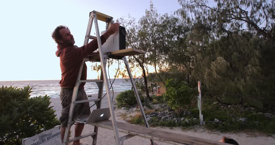 A man climbing a ladder near a beach and adjusting a camera that is balanced on it