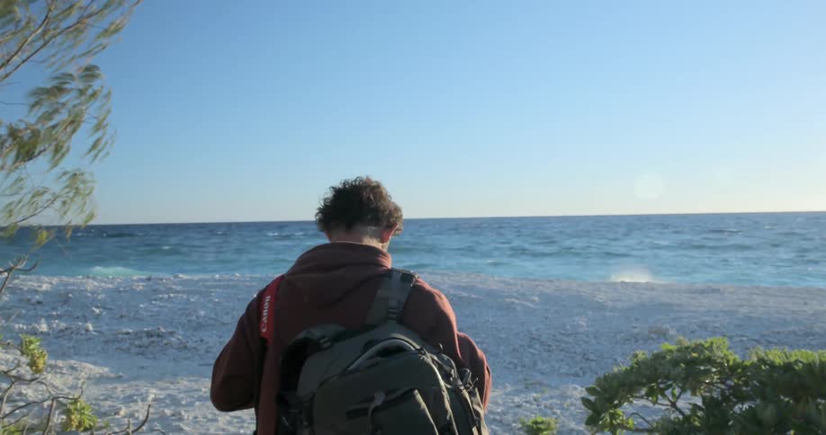 A man walks towards the water holding a camera camera and backpack