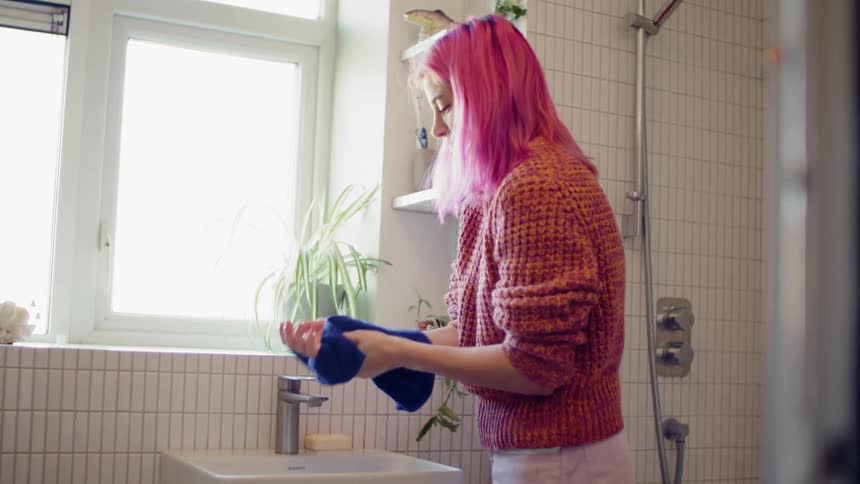 A teenage girl with pink hair washes her hands in a bathroom sink and dries them on a hand towel