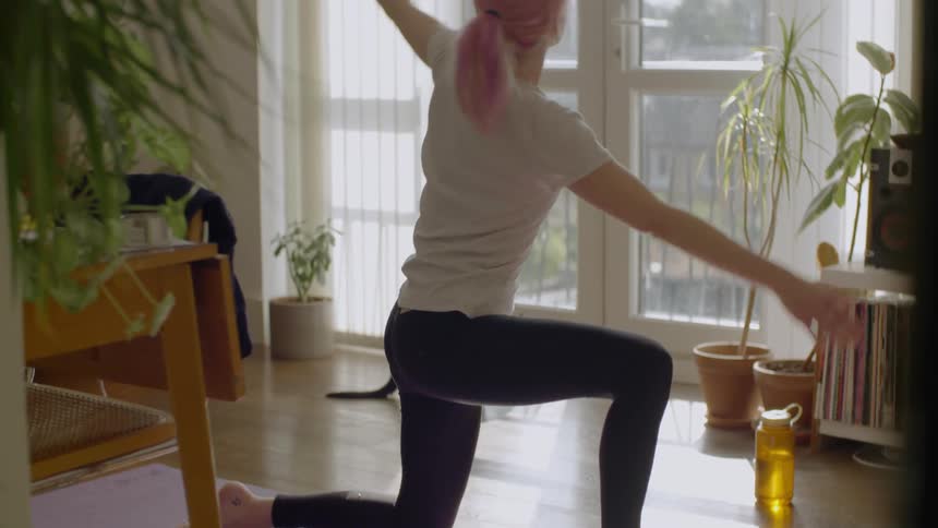 A pink haired woman performing stretches in her living room